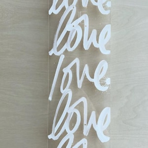 May include: Clear acrylic skateboard deck with the word "love" repeated in white brushstrokes. The word is written in a cursive style. The word "blacklist" is written in black at the bottom.