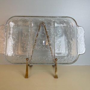 May include: A clear, rectangular glass serving dish with three compartments, featuring a floral pattern. The dish is supported by a gold-colored metal stand. The serving dish has handles on each side.