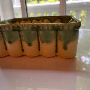 May include: A rectangular ceramic planter with a scalloped edge. The planter has a two-tone color scheme, with a creamy yellow base and green dripping glaze. The planter is sitting on a white surface.
