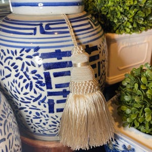 May include: A ceramic ginger jar featuring a white base with blue hand-painted patterns and Chinese characters. A long, cream-coloured decorative tassel with a multi-layered knotted top and a full, flowing fringe hangs from the jar's lid.