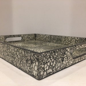 May include: A square decorative tray with a black and white speckled pattern. The tray has a black border and cut-out handles on each side. The tray is designed for serving or display.