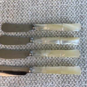 May include: Four butter knives with stainless steel blades and mother-of-pearl handles. The handles have a bamboo-like design. The knives are arranged horizontally on a textured, light gray surface.