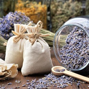 Può includere: Due sacchetti di lino beige legati con spago, pieni di boccioli di lavanda, sono posti davanti a un barattolo di vetro traboccante di lavanda essiccata. Un cucchiaio di legno è appoggiato vicino, circondato da lavanda sparsa.