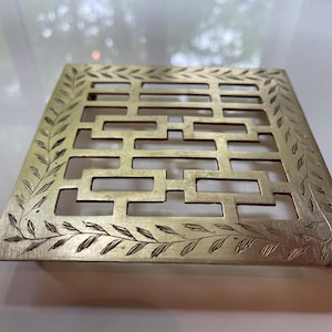 Peut inclure: Un couvercle de ventilation décoratif carré de couleur laiton avec un motif géométrique de découpes rectangulaires. La bordure présente un motif de feuilles gravées. Le couvercle de ventilation est probablement pour la décoration intérieure.
