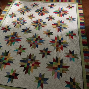 May include: A white quilt with a patchwork design featuring a variety of colors and patterns. The quilt is made up of multiple star-shaped blocks, each with a different color combination. The quilt is bordered with a green and white striped fabric.