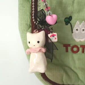 May include: A light green corduroy bag featuring a white cat charm with a pink collar, a black ribbon, a pink heart bead, and clear iridescent beads on a silver ball chain. The bag has brown trim and an embroidered white creature with the text TOT.