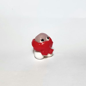May include: A small, rounded figurine featuring a smooth, light purple head with two black circular eyes, wrapped in a thick, bright red scarf. The base of the figure is white and rounded, creating a compact, smooth-textured character design.