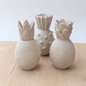 Può includere: Tre vasi in ceramica color bianco sporco a forma di ananas. I due vasi esterni presentano cime appuntite simili a foglie e corpi lisci, mentre il vaso centrale ha una cima strutturata e un corpo coperto da piccoli rilievi circolari.