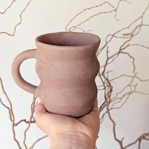 May include: A handmade, light brown ceramic mug with a unique, textured design. The mug has a rounded body with a handle and is held in a person's hand. The mug is approximately 4 inches tall and is a piece of pottery.