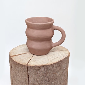 May include: A handmade, terracotta-colored ceramic mug with a unique, rounded design. The mug features a handle and sits on a wooden tree stump, showcasing its earthy tones and artisanal craftsmanship. The mug's dimensions are not visible.