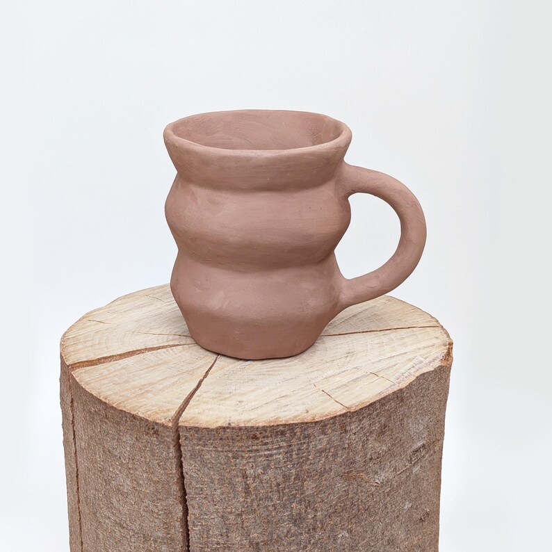 May include: A handmade, terracotta-colored ceramic mug with a unique, rounded design. The mug features a handle and sits on a wooden tree stump, showcasing its earthy tones and artisanal craftsmanship. The mug's dimensions are not visible.