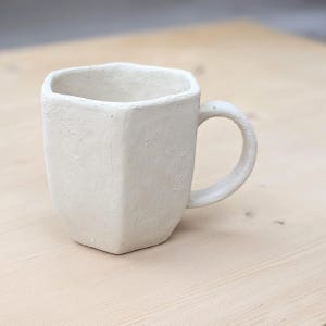 Może przedstawiać: Ręcznie wykonany, kremowy ceramiczny kubek o geometrycznym, fasetowanym wzorze. Kubek ma zaokrąglony uchwyt i teksturowaną powierzchnię. Kubek stoi na jasnej drewnianej powierzchni. Ten wyjątkowy kubek jest idealny do kawy lub herbaty.