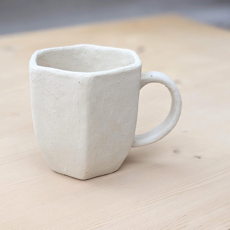 Może przedstawiać: Ręcznie wykonany, kremowy ceramiczny kubek o geometrycznym, fasetowanym wzorze. Kubek ma zaokrąglony uchwyt i teksturowaną powierzchnię. Kubek stoi na jasnej drewnianej powierzchni. Ten wyjątkowy kubek jest idealny do kawy lub herbaty.