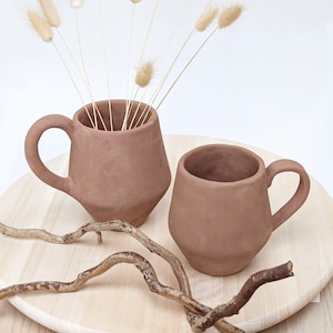 May include: Two terracotta-colored ceramic mugs with rounded handles sit on a light wood surface. One mug holds dried beige flowers. A curved, brown twig rests near the mugs, adding a natural element to the composition. The mugs have a rustic, handmade appearance.
