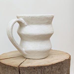 May include: A handmade white ceramic mug with a textured surface and a curved handle. The mug has a unique, ridged design and sits on a natural wood slice. This handcrafted mug is perfect for coffee or tea.