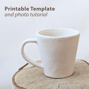 Op de afbeelding: Een witte keramische mok met een getextureerd oppervlak en een gebogen handvat. De mok staat op een houten oppervlak. De afbeelding bevat de tekst "Printable Template and photo tutorial".