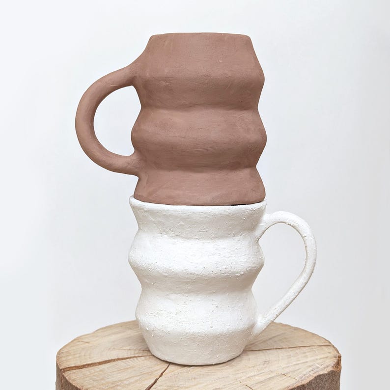 May include: Two handmade ceramic mugs stacked on a wooden surface. The bottom mug is white, and the top mug is a muted brown. Both mugs have a unique, ridged design and curved handles, showcasing a modern, artistic aesthetic.