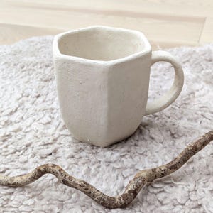 Peut inclure: Une tasse en céramique blanc cassé faite à la main, avec un design géométrique. La tasse a une forme angulaire unique et une anse arrondie. Elle est posée sur une surface blanche et duveteuse, avec une branche brune texturée.