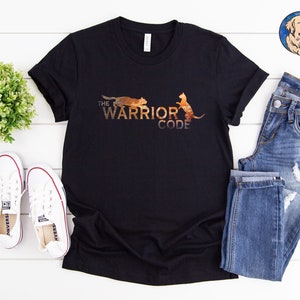 May include: Black t-shirt with the text "THE WARRIOR CODE" and a graphic of two cats in shades of brown and orange. The shirt is displayed with blue jeans and white sneakers.