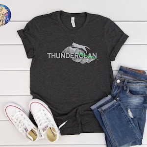 May include: A dark gray t-shirt with a white graphic of a cat jumping through a cloud with the text "THUNDERCLAN". The shirt is paired with blue jeans and white sneakers.