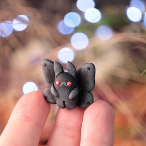 May include: A small, dark gray clay figurine of a moth-like creature with red eyes and a small mustache. The figurine has large, textured wings and rabbit-like ears. The background is blurred with bokeh lights.