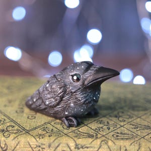 May include: A small, black raven figurine with white speckles, perched on a patterned surface. The raven has a detailed, textured appearance and a glossy eye. The background features soft, out-of-focus lights, creating a whimsical effect.