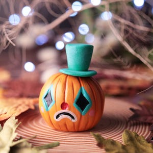 Könnte beinhalten: Eine orangefarbene Kürbisfigur mit einem traurigen Clowngesicht, die einen türkisfarbenen Zylinder trägt. Der Kürbis hat schwarze rautenförmige Augen mit türkisfarbenen Umrissen, eine rote Nase und rosa Wangen. Verschwommener Hintergrund mit Herbstblättern.