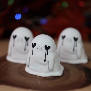 May include: Three white ghost figurines with black heart-shaped eyes and tear streaks. The ghosts have a rounded shape and sit on a wooden surface. The background is blurred with bokeh lights.