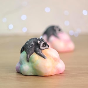 May include: A small figurine of a raccoon with dark gray fur patterned with white stars, resting on top of a rounded, multi-colored cloud shape featuring blended pastel shades of pink, yellow, green, and blue. A second identical figurine is visible behind.