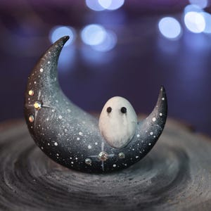 May include: A small white ghost figurine with black eyes sits on a black crescent moon with white stars and glitter. The moon is on a dark wood surface.