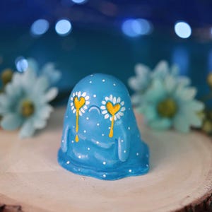 May include: A blue blob-shaped figurine featuring white polka dots and two eyes depicted as yellow hearts surrounded by white daisy petals. The figure has a small curved line for a mouth and appears to have yellow liquid dripping from its eyes.