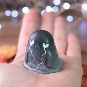 May include: A small, dark gray figurine shaped like a blob with white heart-shaped eyes and tears. The figurine has a cracked design and is held in an open palm. The background is blurred with bokeh lights.