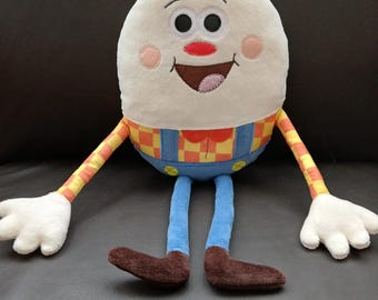 Op de afbeelding: Een eivormige knuffel met een wit gezicht, zwarte ogen, een rode neus en een brede glimlachende mond. Het draagt een blauwe tuinbroek over een geel en oranje geblokt hemd, met lange dunne armen, witte handen, blauwe benen en bruine voeten.