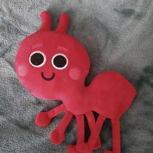 May include: A red plush ant toy with big black eyes and a smiling mouth. The ant has six legs and is lying on a gray fuzzy surface.