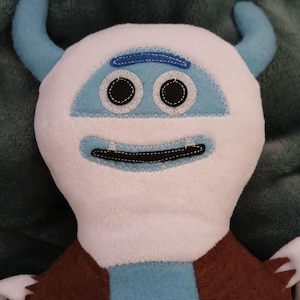 May include: A white plush toy with blue accents, resembling a monster. It has two large black eyes, a stitched-on mouth, and two blue horns. The toy is wearing a brown vest.