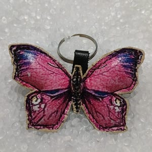 May include: A butterfly-shaped keychain with a pink and purple design. The wings have a detailed pattern, and the keychain has a silver ring and a black leather strap. The butterfly is made of a textured material.