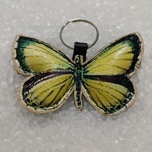 Könnte beinhalten: Ein gelber, schmetterlingsförmiger Leder-Schlüsselanhänger mit grünen und schwarzen Akzenten. Der Schlüsselanhänger hat einen silbernen Metallring.