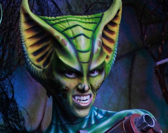 May include: A green and yellow alien creature with large ears and sharp teeth. The creature is wearing a green and yellow costume and is holding a gun.