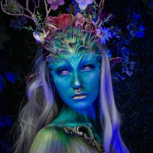 May include: A woman with blue and green face paint and a floral crown. Her eyes are bright blue and she has a silver nose ring. Her hair is long and white.