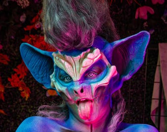May include: A person wearing a blue and white bat costume with a pink tongue sticking out. The costume has a detailed face with sharp teeth and large ears. The person's hair is styled in a spiky, purple and grey mohawk.
