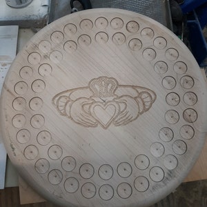 May include: A wooden round game board with a Celtic knot design featuring a heart in the center. The board has 60 holes around the edge.