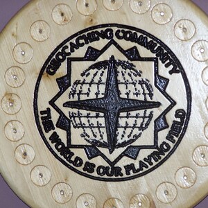 May include: A wooden round plaque with a black and white design of a compass and the text "Geocaching Community The World Is Our Playing Field". The plaque has 24 small holes around the edge.