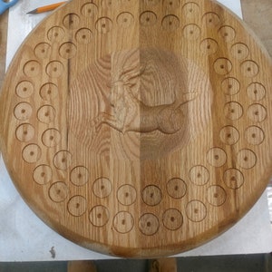 May include: A wooden game board with a carved rabbit design in the center. The board has a circular shape and is surrounded by a ring of evenly spaced holes.