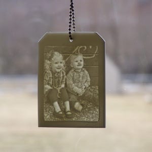 May include: A rectangular, olive-toned car charm with a chain. The charm features a sepia-toned image of two children sitting together. The image is etched into the surface, creating a vintage aesthetic. The charm hangs against a blurred background.