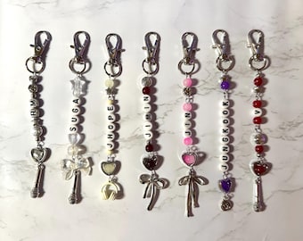 May include: A collection of seven keychains with silver-tone clasps. Each keychain features a beaded strand with a name spelled out in white letter beads. Charms include hearts, bows, and microphones. The names are RM, SUGA, JHOPE, JIMIN, JIN, JUNGKOOK, and V.