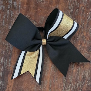 May include: A large hair bow featuring black fabric with layered accents of white and gold glitter ribbon. The bow is constructed with a central gold glitter knot and two long, pointed tails, each decorated with a vertical stripe of gold glitter.