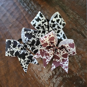 May include: Three cow print hair bows in black and white, black and brown, and white and brown.