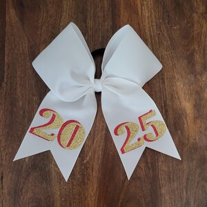 May include: A white hair bow with red and gold glitter accents. The bow features the numbers "2025" in a decorative font. The bow is made of a sturdy material and is likely used for cheerleading or other sports.