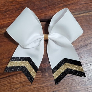 May include: A large white hair bow made of grosgrain ribbon, featuring a central gold glitter knot. The two bottom tails are decorated with a chevron pattern consisting of alternating black and gold glitter stripes on a white background.