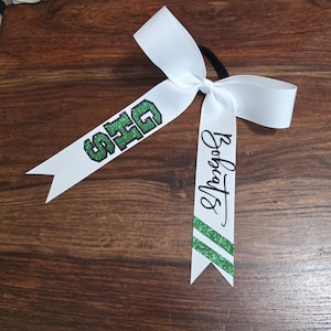 May include: A white hair bow attached to a black elastic band. One ribbon tail features the green glitter text GHS, while the other tail displays the black script text Bobcats above two diagonal green glitter stripes at the bottom edge.